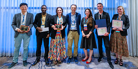 Seven individuals pose together. Multiple individuals are either holding an award plaque, certificate, or envelope. 