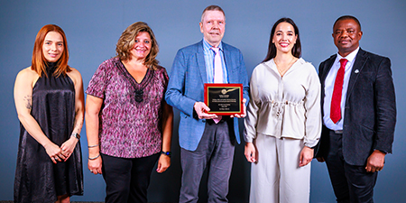 Five individuals pose together, with the center individual holding an award plaque. 