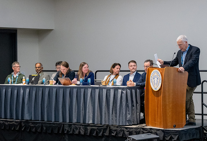 Eight individuals sit behind a long, curtained table, while a ninth stands behind a podium reading aloud from some notes. 