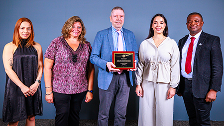 Five individuals pose together, with the center individual holding an award plaque. 