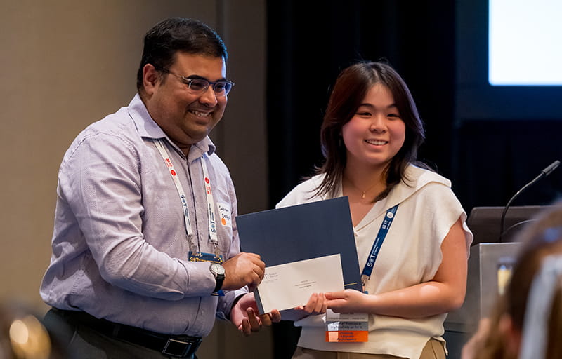Two individuals are standing next to each other and smiling for an unseen camera. Between them, they are holding a certificate and envelope.