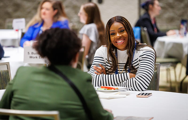  An individual sits with their back to the camera. Across them at a table sits another individual who is smiling. In the background, you can see other individuals chatting at tables.