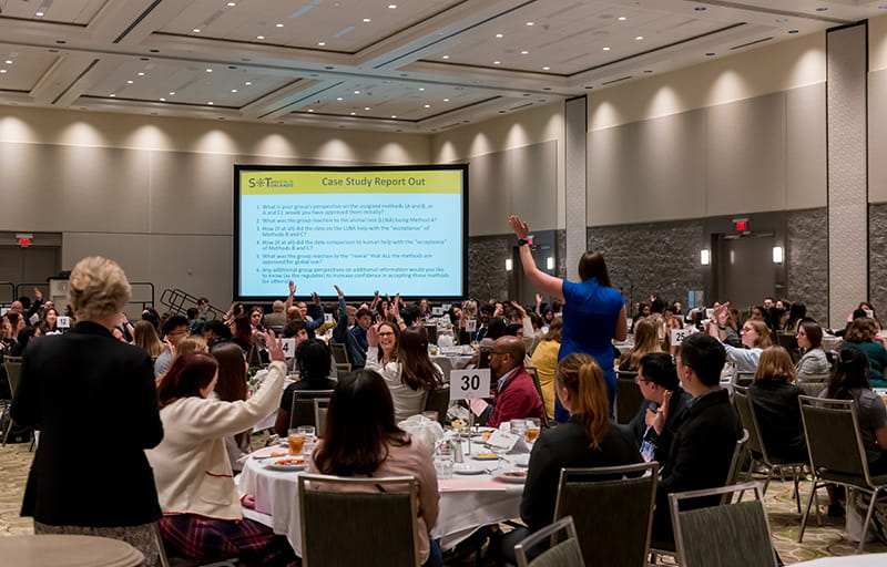 A room is full of large, round tables. Individuals are seated at the tables. Some are raising their hands. In the distance, you can see a projection screen.