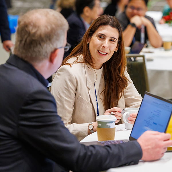 Two individuals sit at a table chatting. One has their back to the camera. The other individual is smiling.