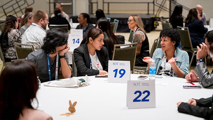 Individuals are sitting around a large, round table and are chatting. You can see other individuals in the background employed in the same activity. 