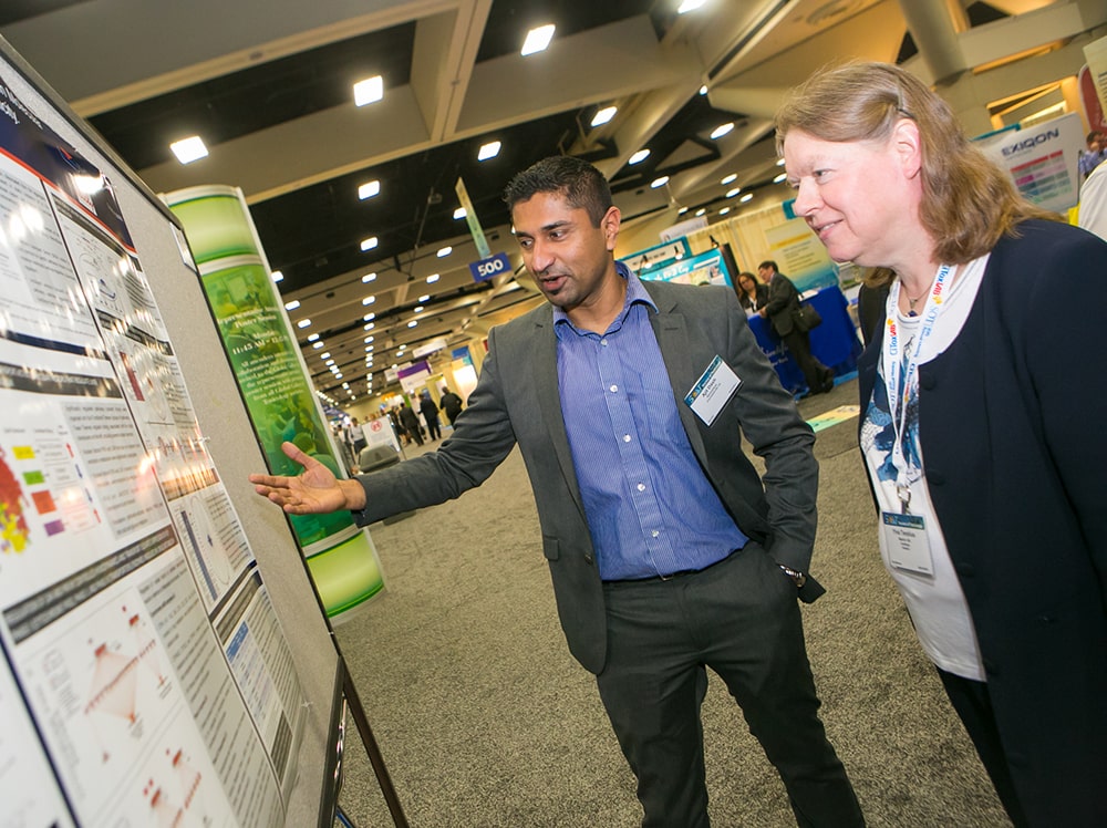 To the left is a poster mounted on a large gray board. Toward the middle of the photo, a man stands looking at and gesturing to the poster. Next to him, on the right-hand side of the photo, stands a woman who also is looking at the poster.