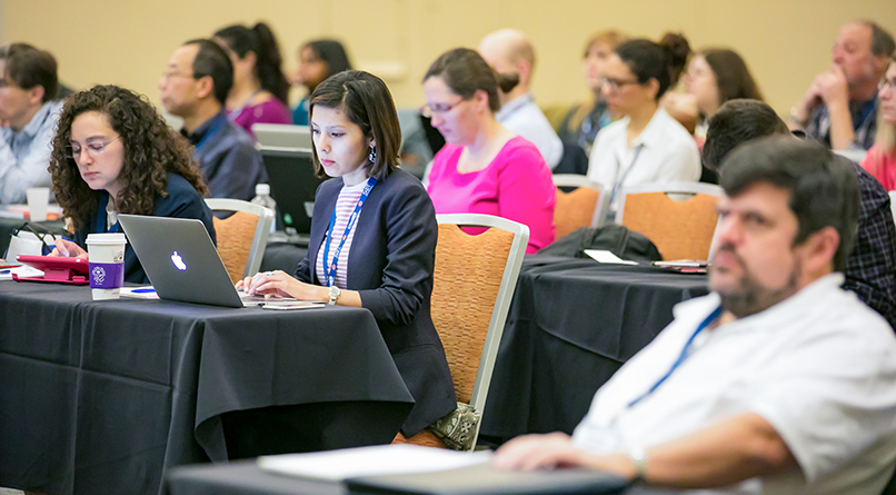 Multiple rows of long tables covered with black tableclothes are visible. Individuals are seated behind each table so that they all are facing the same direction. In focus in the middle of the image is a young woman looking at the laptop that is sitting on the table in front of her.