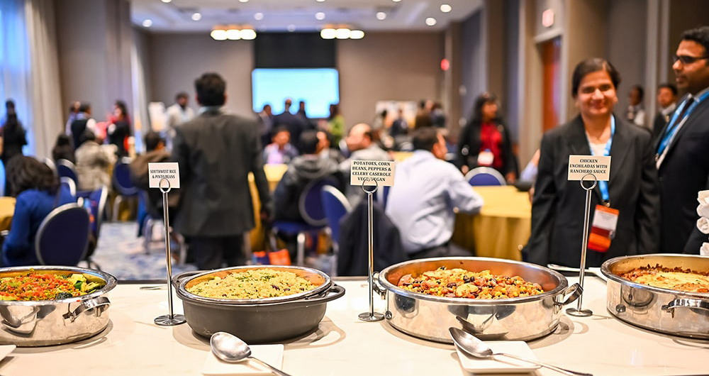 In the foreground is a table holding four serving pots. Serving spoons are resting in front of the pots, while a metal stand sits next to each pot. The metal stand holds an index card detailing the dish in the pot. In the background, out of focus, people are milling and sitting at tables.