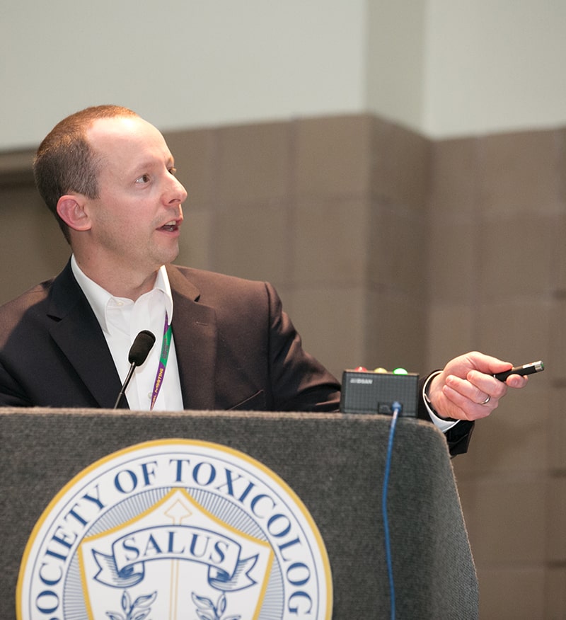 A man stands behind a lectern. He is looking to his left. His left hand is raised, holding a laser pointer, and turned toward the direction in which he is looking. He is in mid-speech.
