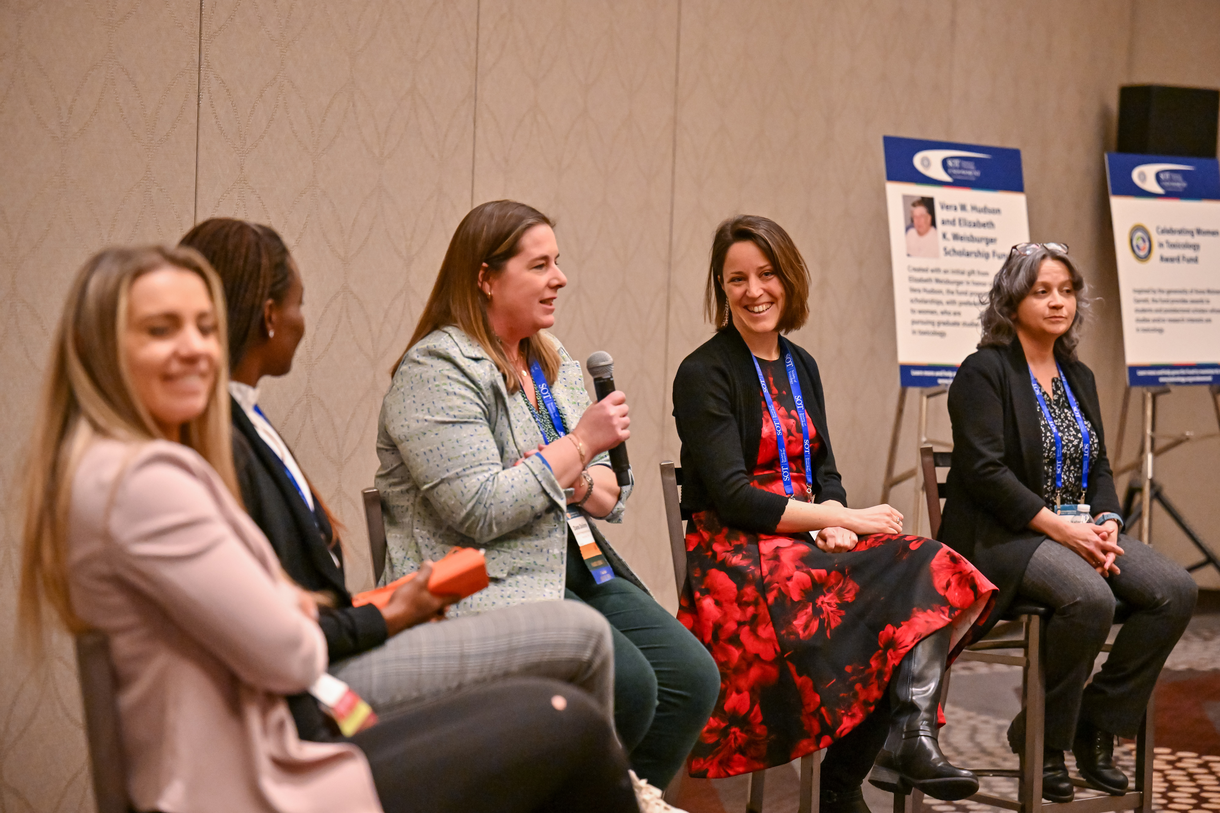 Five individuals sit on stools that form an arch shape. The individual in the middle is holding a microphone and speaking.
