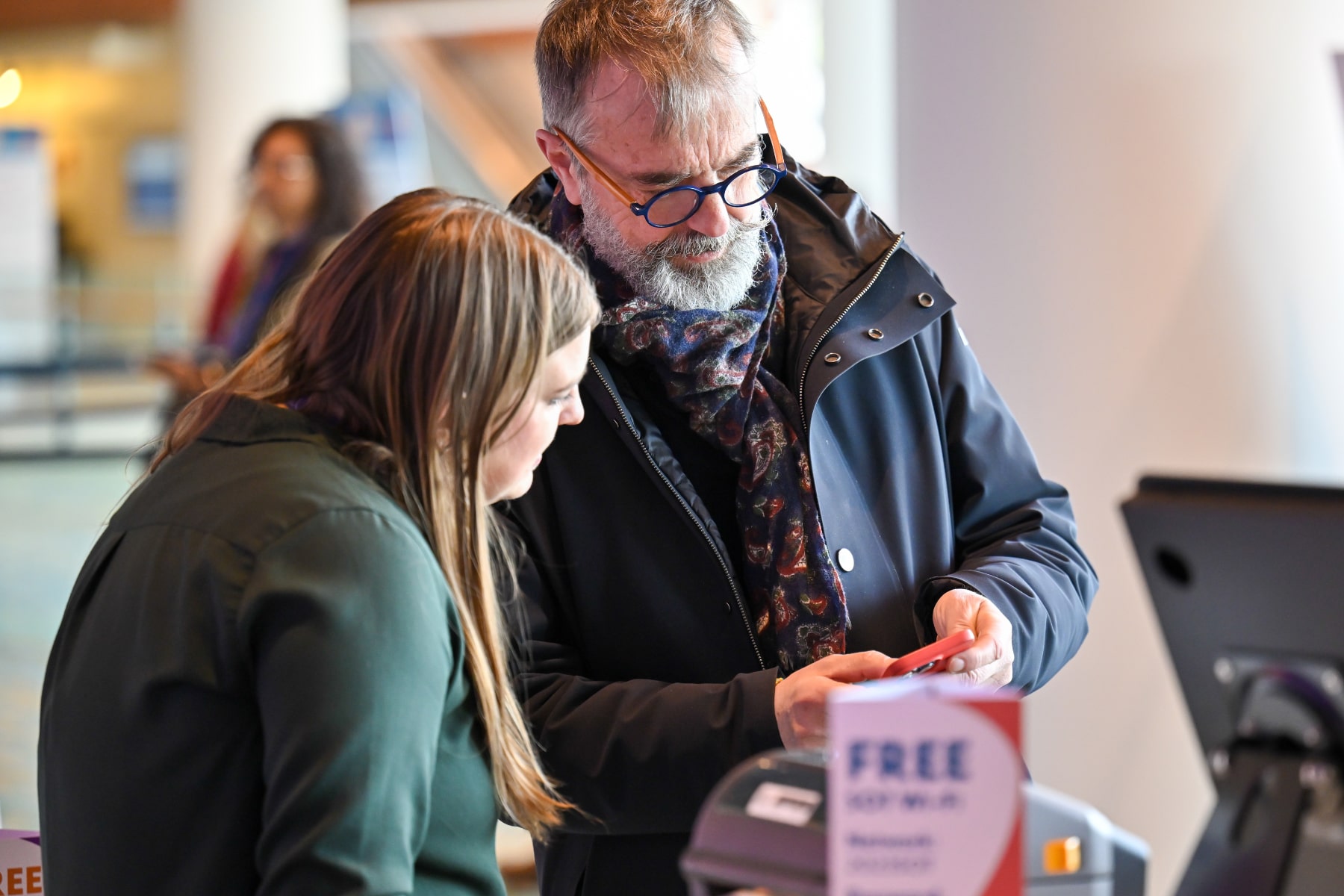 Two individuals stand together. One is holding a phone and looking at it, while the second individual is leaned toward the phone also looking at it.