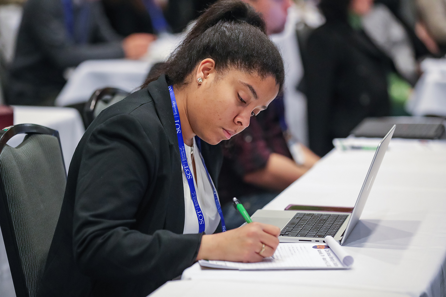 An individual sits at a table with an open laptop in front of them. They are writing in a notepad that sits to the left of the laptop