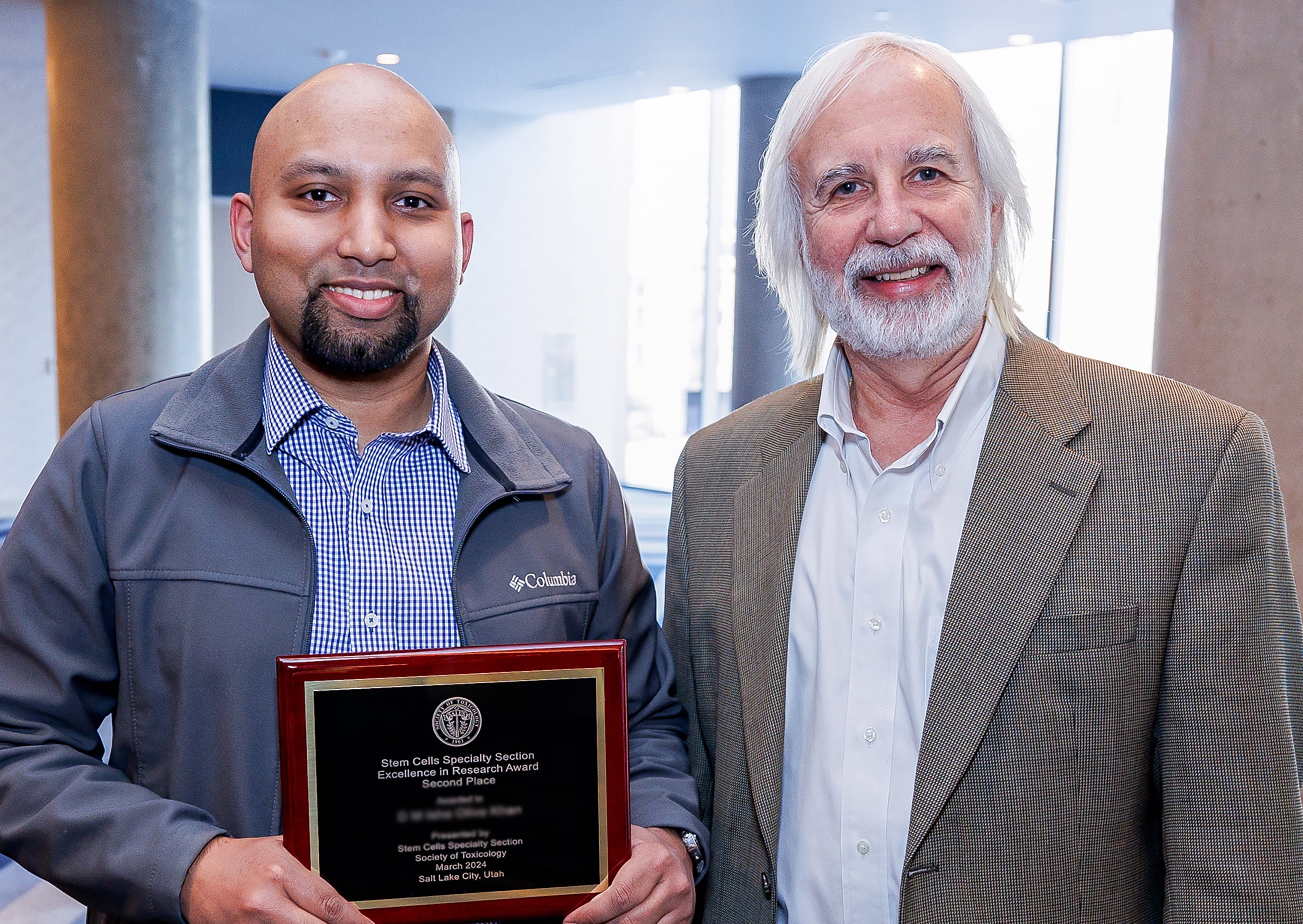 Two individuals stand next to each other smiling at the camera. One individual is holding an award plaque.