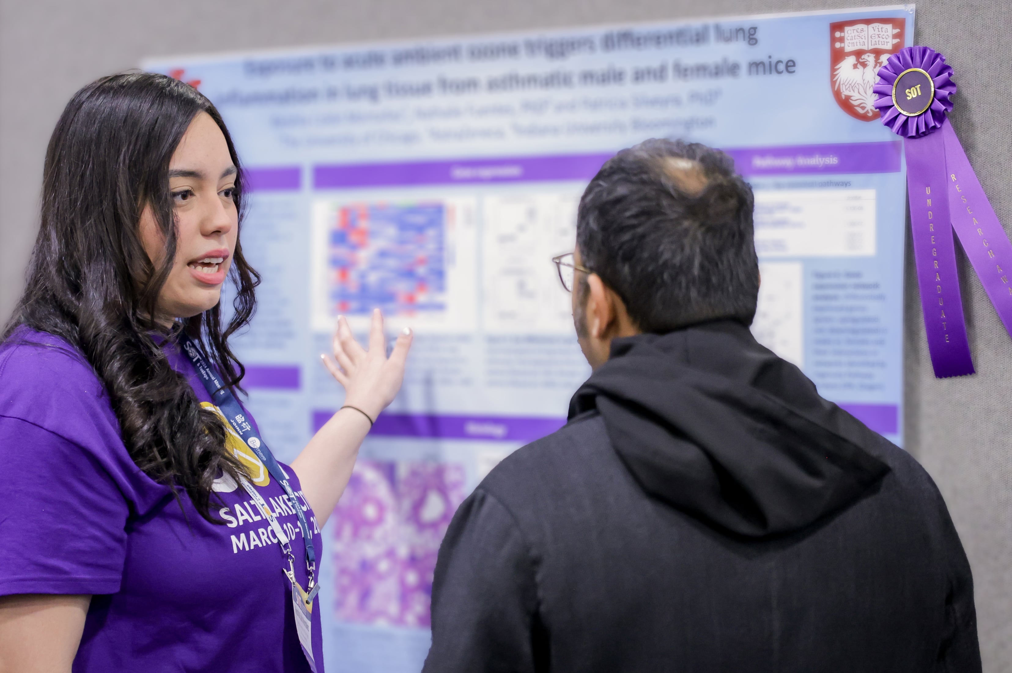 An individual stands in front of a mounted posterboard. They are turned sideways next to the poster speaking with an individual who is looking at the poster. Tacked next to the poster is a purple award ribbon.