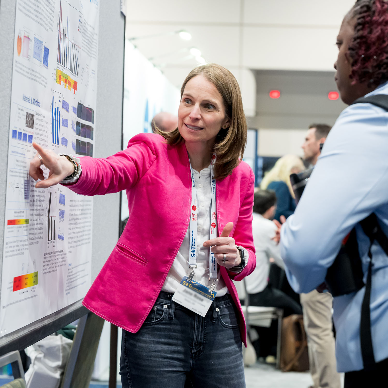 An individual is pointing toward a poster board. Another individual stands next to them, listening to what they are saying.