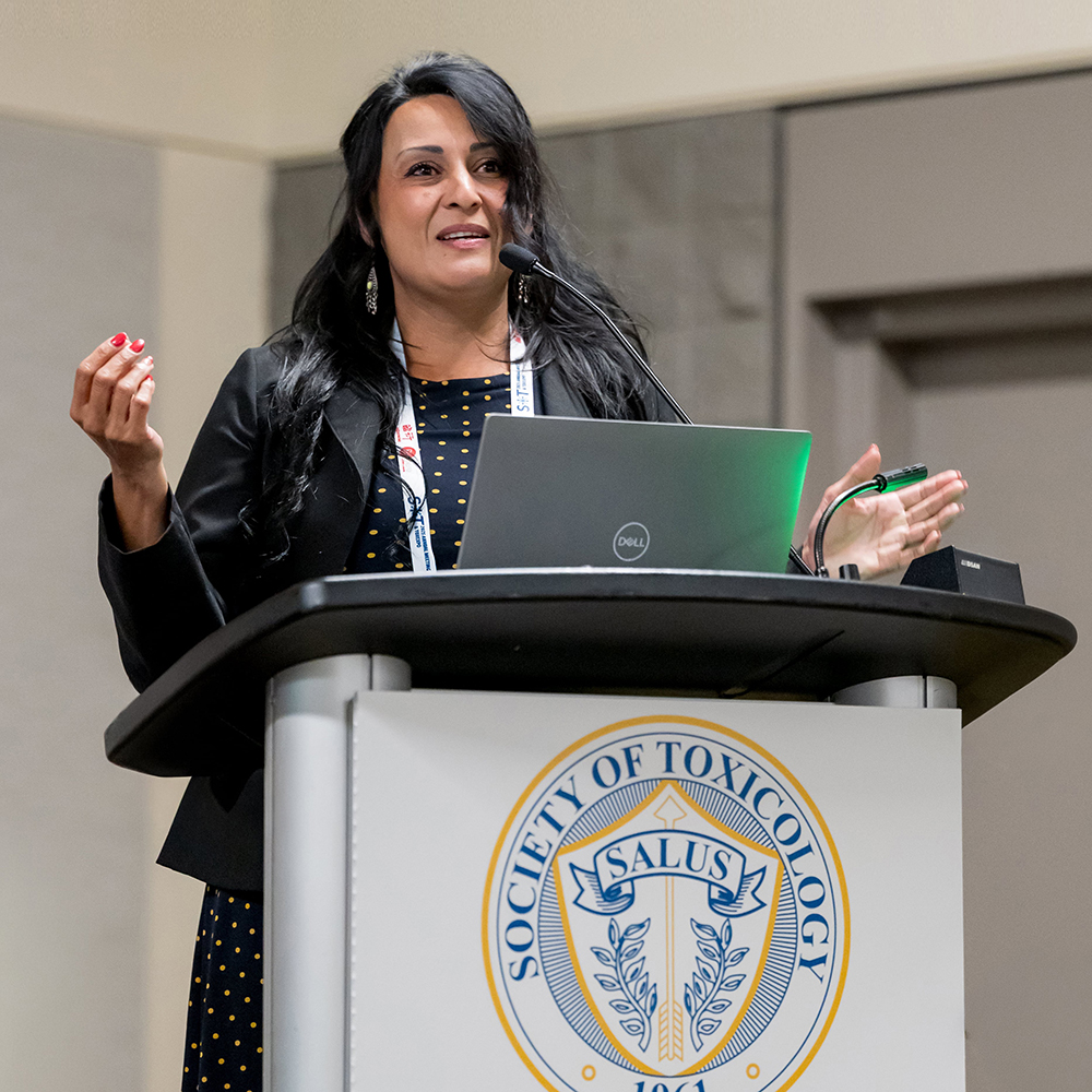 An individual stands behind a lectern that has the SOT salus on its front. The individual is speaking and gesturing.