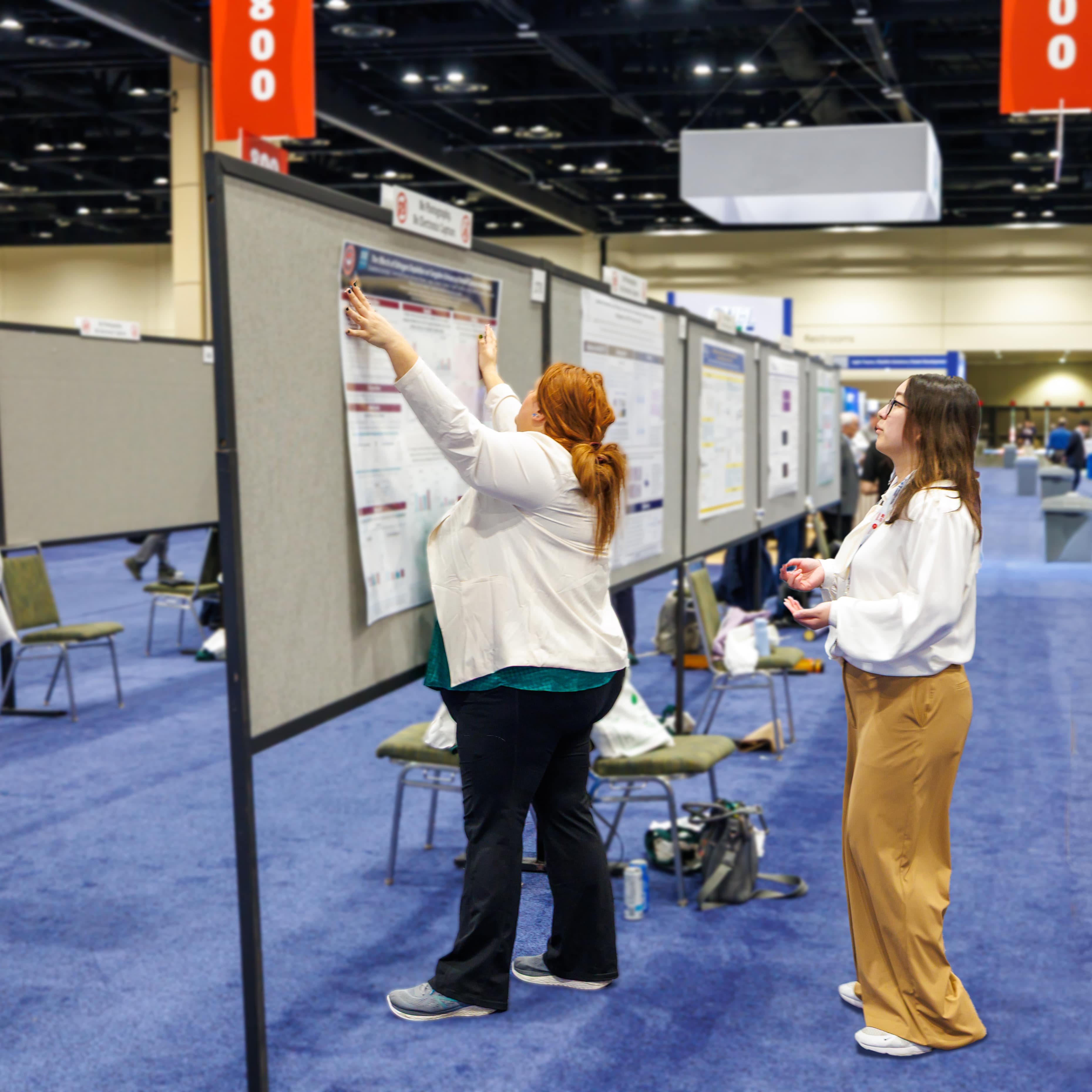An individual is holding a poster up to a gray display board while another individual looks on with pushpins in their hand.