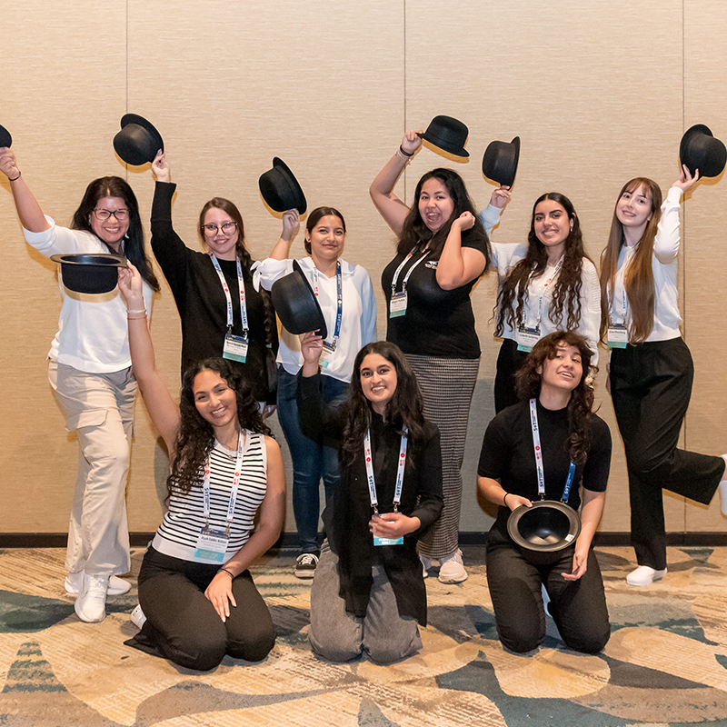 A diverse group of young individuals are posed for the camera. All the individuals are wearing black bowler hats and are smiling.