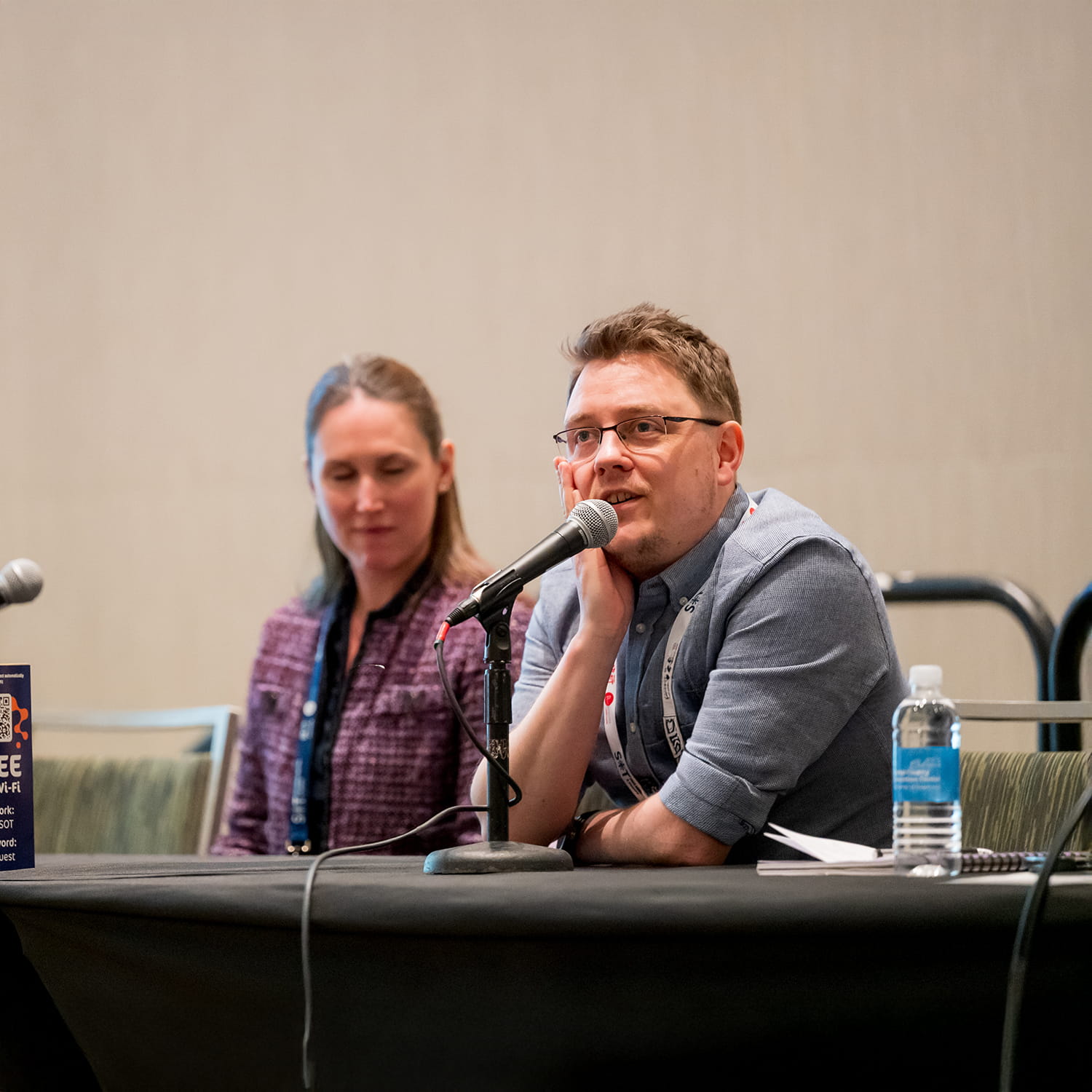 Two individuals sit at a table. One of the individuals is speaking into a microphone that is propped on the table.