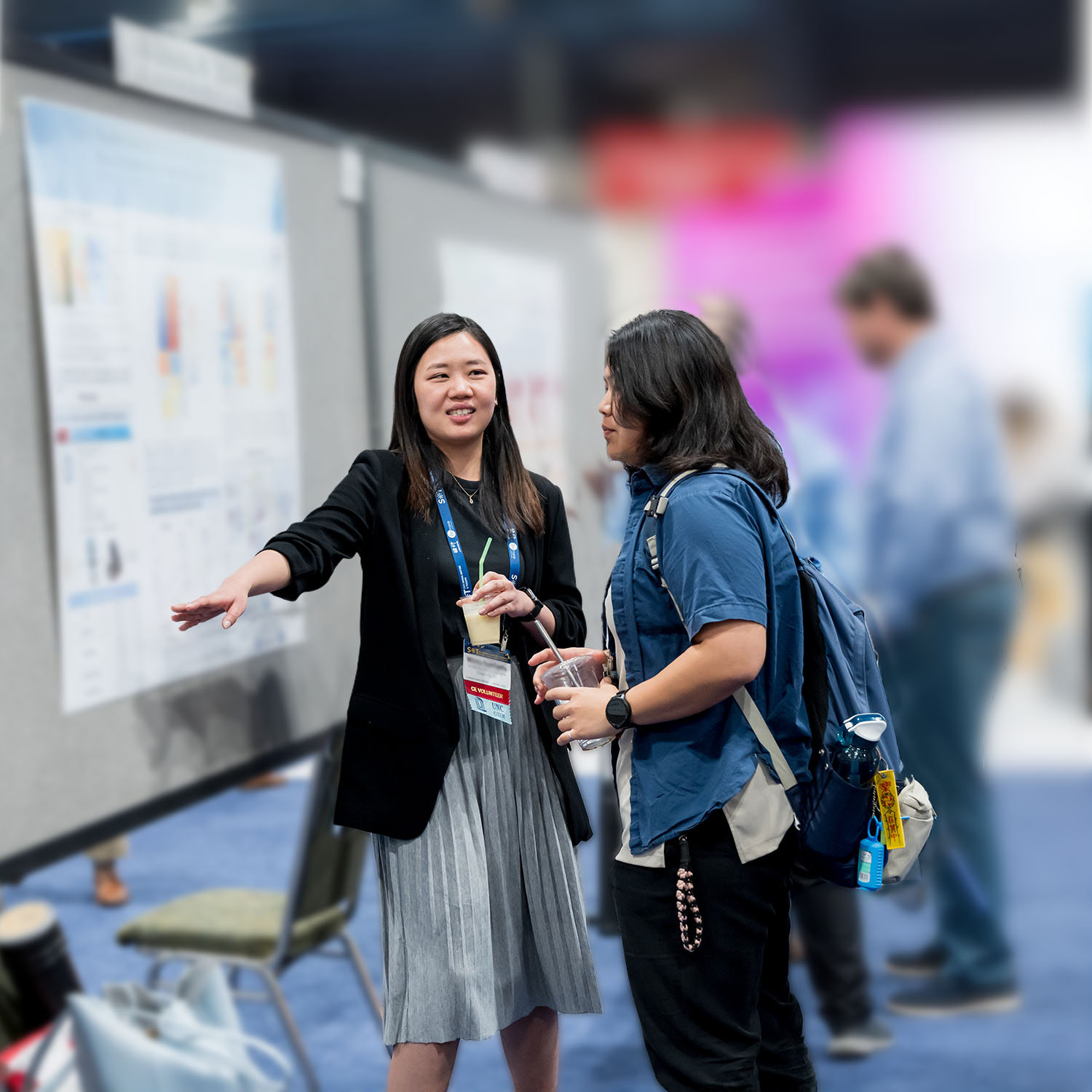 An individual is gesturing toward a poster board while speaking to an individual who is looking at the same poster board.