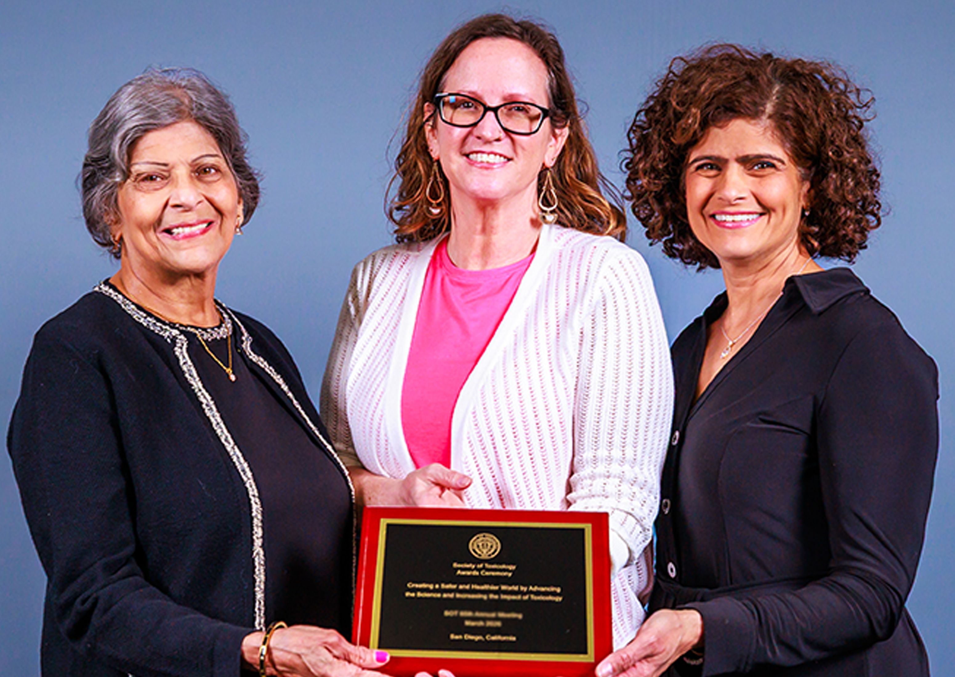 Three SOT Annual Meeting attendees stand together holding an award plaque in front of a blue backdrop. 
