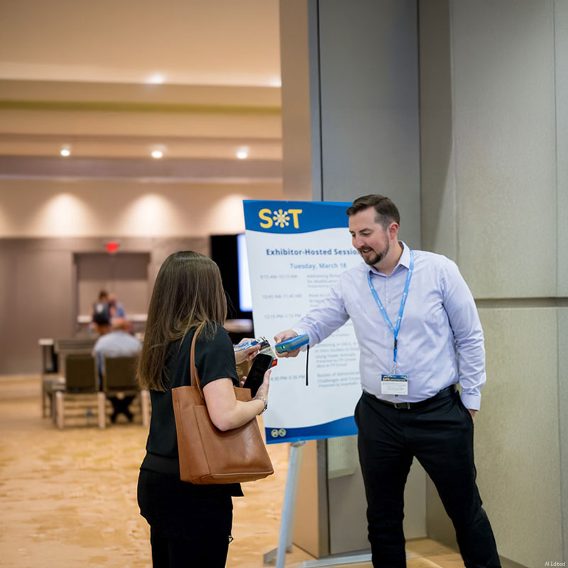 An individual stands next to a sign in front of a doorway. In their hand is an electronic badge scanner. They are in the process of scanning the badge of the individual standing in front of them.