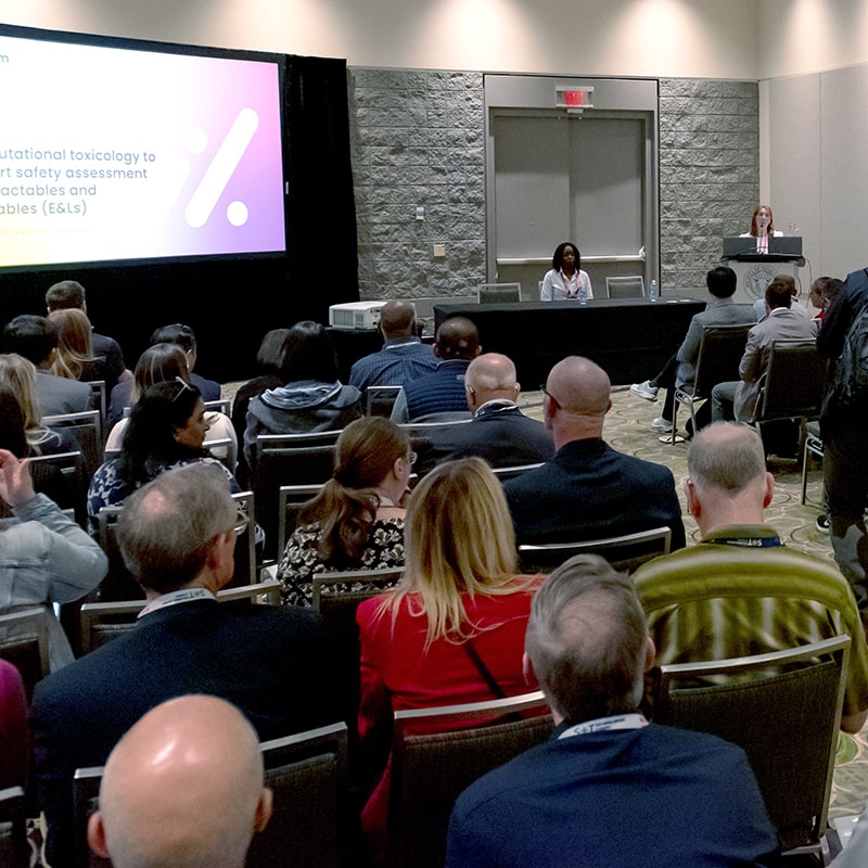 An individual sits at a long, skirted table facing an audience that is seated in rows of chairs with an aisle in the middle. A presentation is being projected on a large screen next to the skirted table.