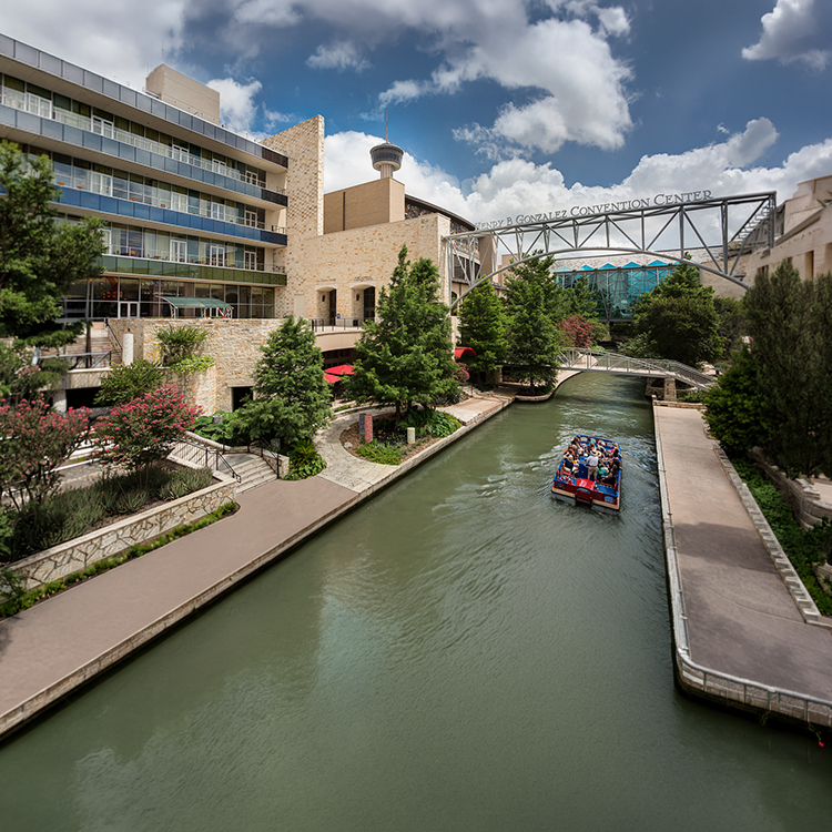 This image shows a buildings on two sides of a waterway with a bridge spanning the waterway. A boat with passengers is visible on the water.