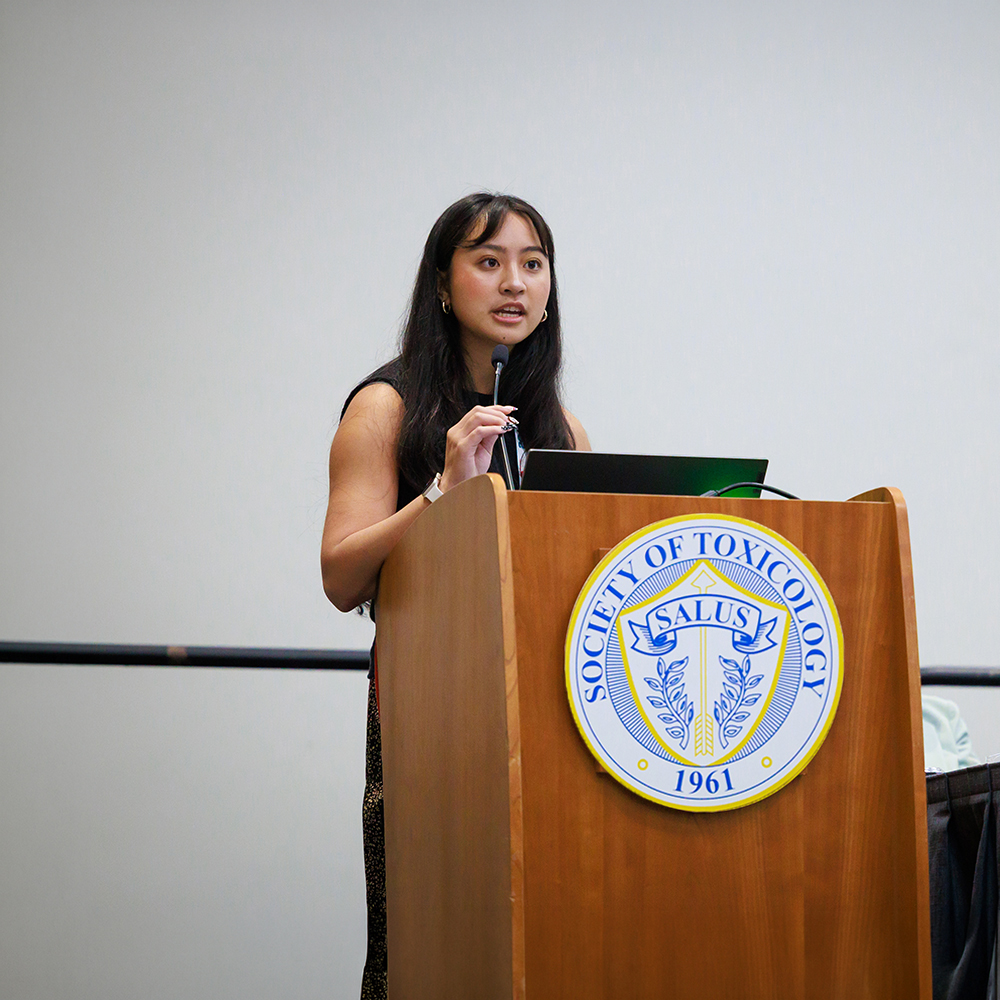 An individual stands behind a wooden podium. The individual is speaking and gesturing.