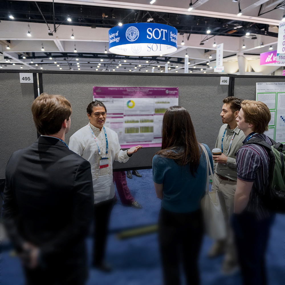 An individual stands in front of a poster display. Four other individuals stand in a semi-circle around the presenter. The presenter is speaking and gesturing.