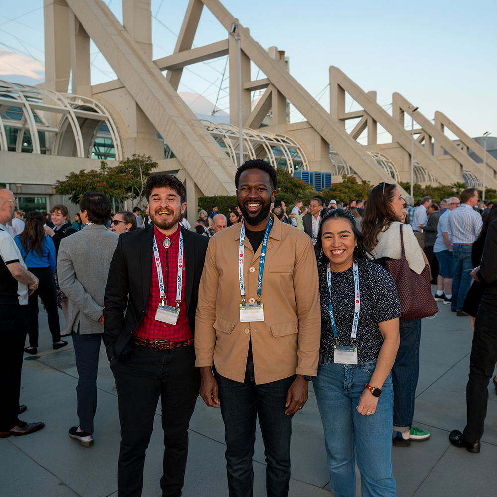 Three individuals stand side-by-side, facing the camera. They are smiling. Behind them, you can see other people gathered and chatting.