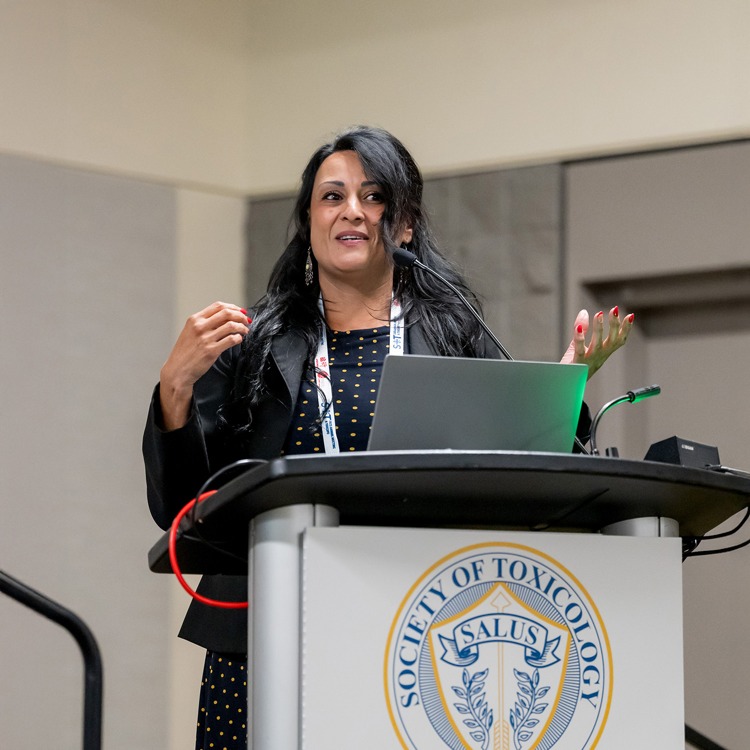 An individual stands behind a podium and is gesturing and speaking. A laptop and microphone are on the podium.