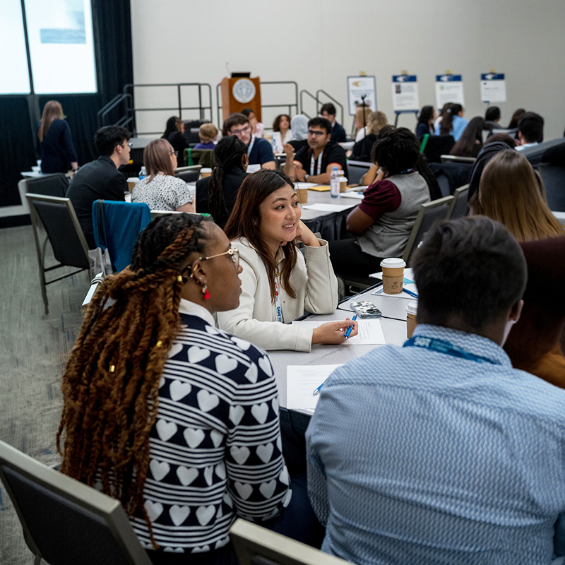 Conference participants seated around tables during a workshop session, listening and discussing in a meeting room.