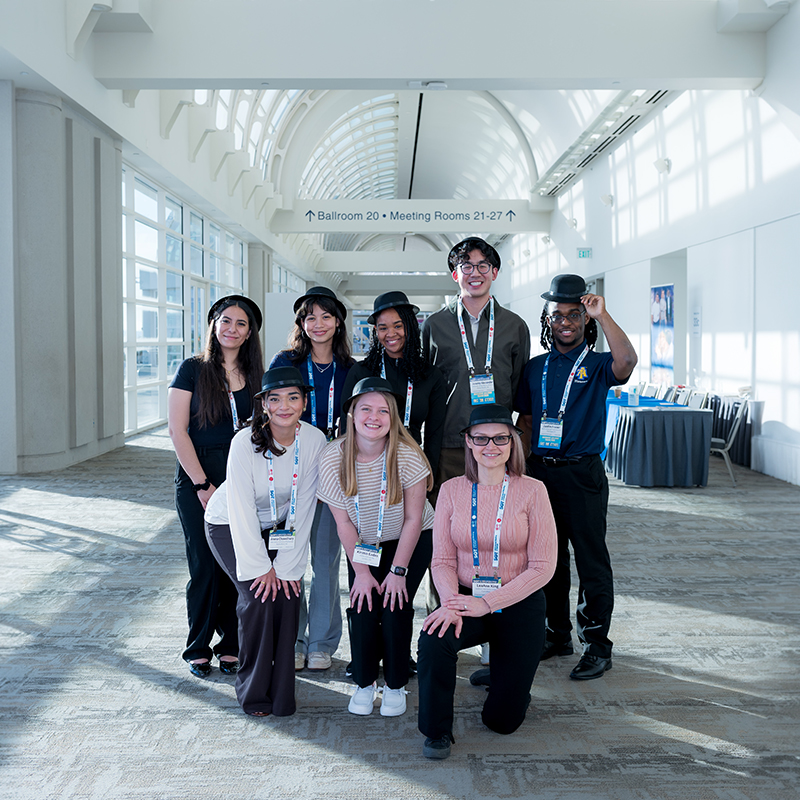 A diverse group of young individuals are posed for the camera. All the individuals are wearing black bowler hats and are smiling.
