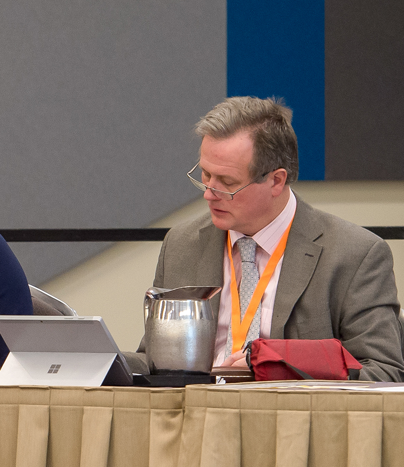 A man in a tie and business jacket sits at a table. He is looking at a tablet that is propped on the table in front of him.