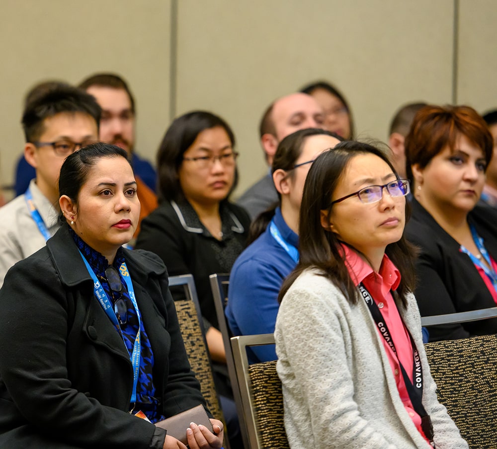 Individuals are seated in rows watching a presentation off-camera. There is not an empty seat visible.