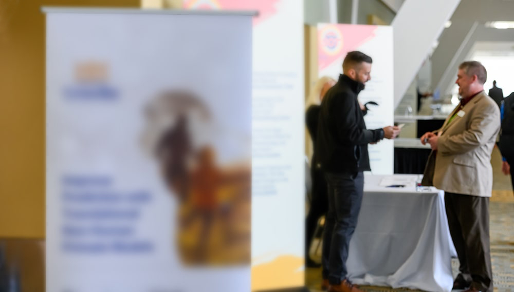 In the left foreground is a promotional sign. In the background to the right of the sign, two men who are out of focus are chatting at a cloth-covered table.