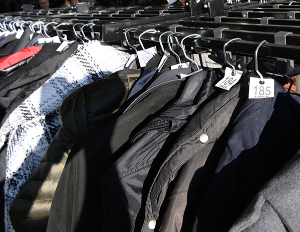 In the foreground, coats are hanging in a long row on a portable metal rack. White tags with numbers are visible on the hangars above the coat collars. Other rows of metal coat racks are visible in the background.