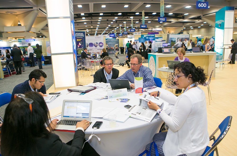 Individuals sit around a round table. On the table are laptops and booklets. All the individuals are busy working on the laptops or looking at the booklets on the table. In the background, you can see the rest of an exhibit hall.