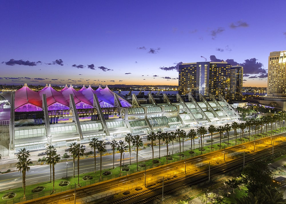 The last remnants of an orange sunset can be seen on the horizon. The image is focused on a large three-story building that covers multiple city blocks. The building is shining from its interior lights since most of its walls are covered with windows and white exterior, while a rooftop feature that looks like a series of ship sails glows bright pink. Lining the street that runs in front of the bulding are palm trees.