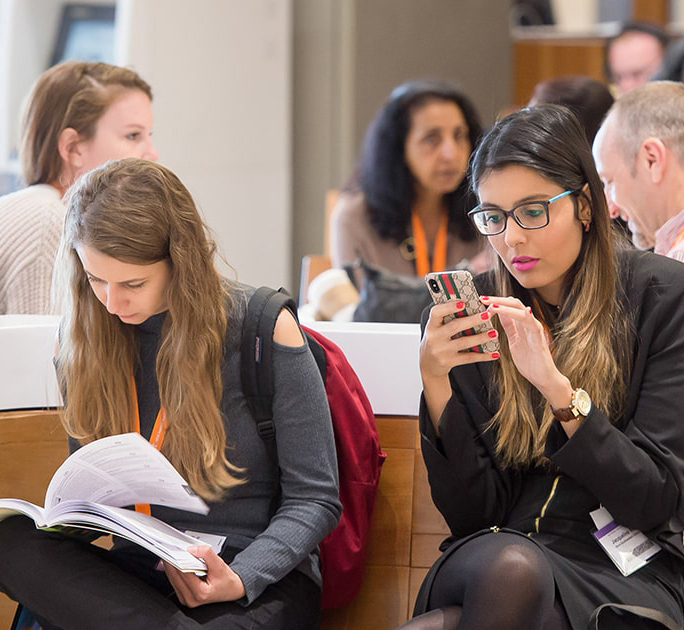 A woman is seated on a bench looking at the mobile phone that she is holding. Other individuals can be seen chatting in small groups behind her.