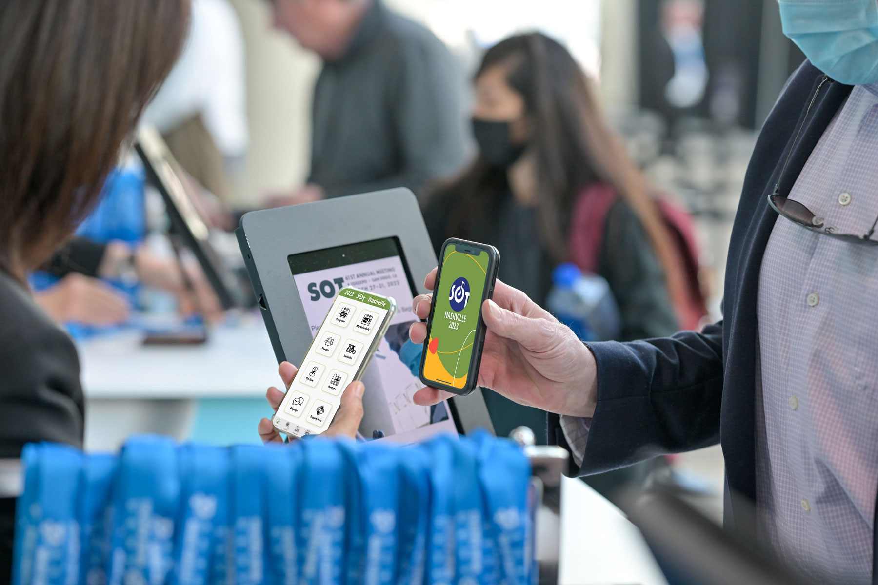 A close up of two people holding phones. One person has the phone angled toward them and is looking at the 2023 SOT Event App home screen. The second person has their phone screen turned toward the first person, with the 2023 SOT Annual Meeting logo and artwork displaying on the screen.