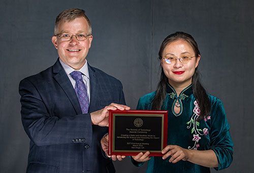 A man and woman stand side by side in front of a gray background. They are posed holding an award plaque between them and are smiling at the camera.