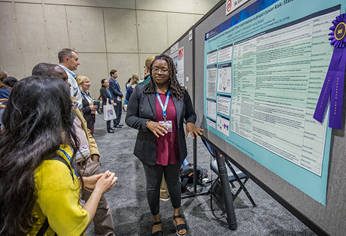A young woman stands next to a poster display. She is looking to her right and speaking to the few individuals that are standing there looking at her poster. Next to the ribbon is a purple rosette ribbon.
