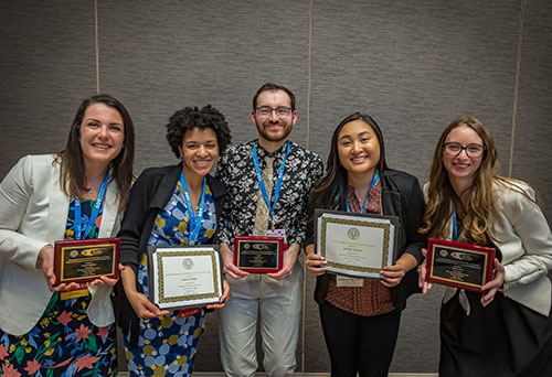 A group of five young people stand side by side but leaning toward each other. They are looking at the camera and smiling. Three of the individuals are holding plaques in front of them, while the other two are holding certificates.
