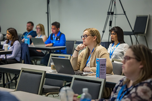In the center of the image is a woman sitting at a long table. She has a laptop in front of her, but she is looking beyond the laptop. Around the woman, you can see other long tables where other people sit with laptops.