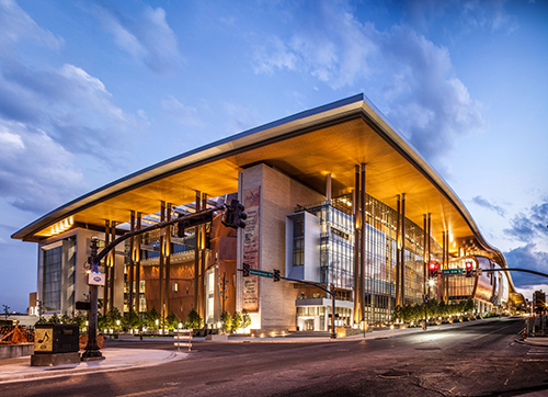 This image of Nashville Music City Center is taken from across the street during the twilight hour. The building is glowing with lights, and you can see the modern architecture full of floor-to-ceiling windows.