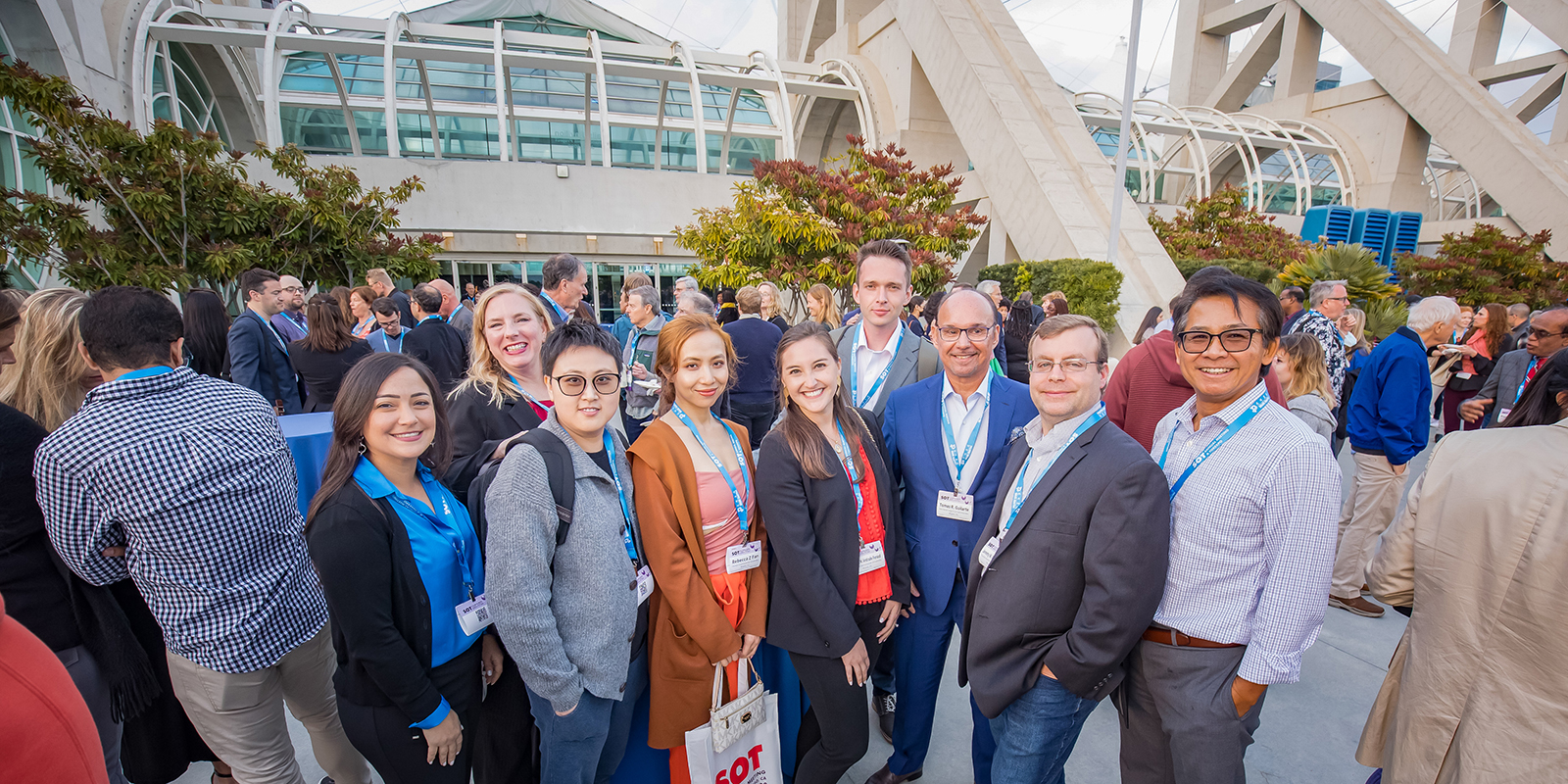 A large group of individuals representing varied ages, genders, and ethnicities, stands posing together. Other individuals are milling in the background.