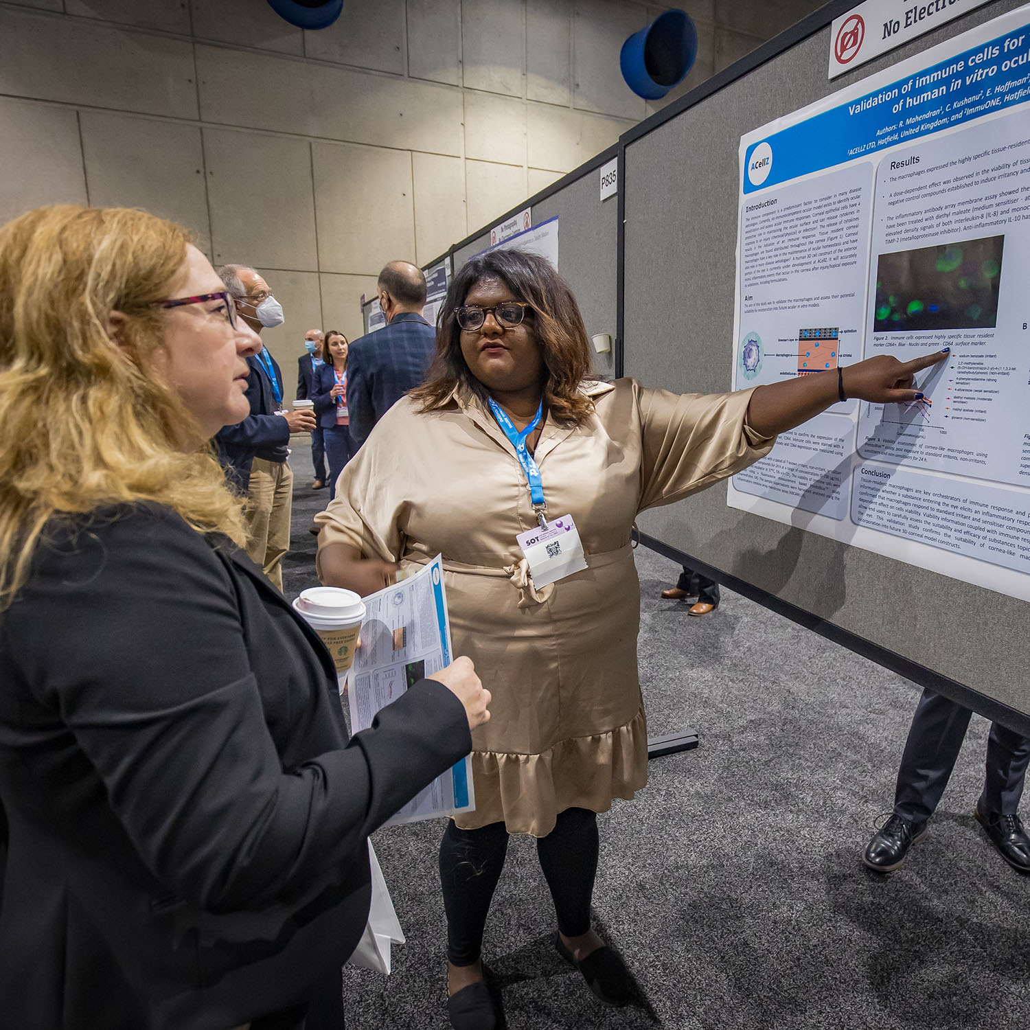 A woman stands next to a large poster. With her left hand, she is pointing to a section of the poster. The woman is looking toward her right where another woman stands looking at the poster.