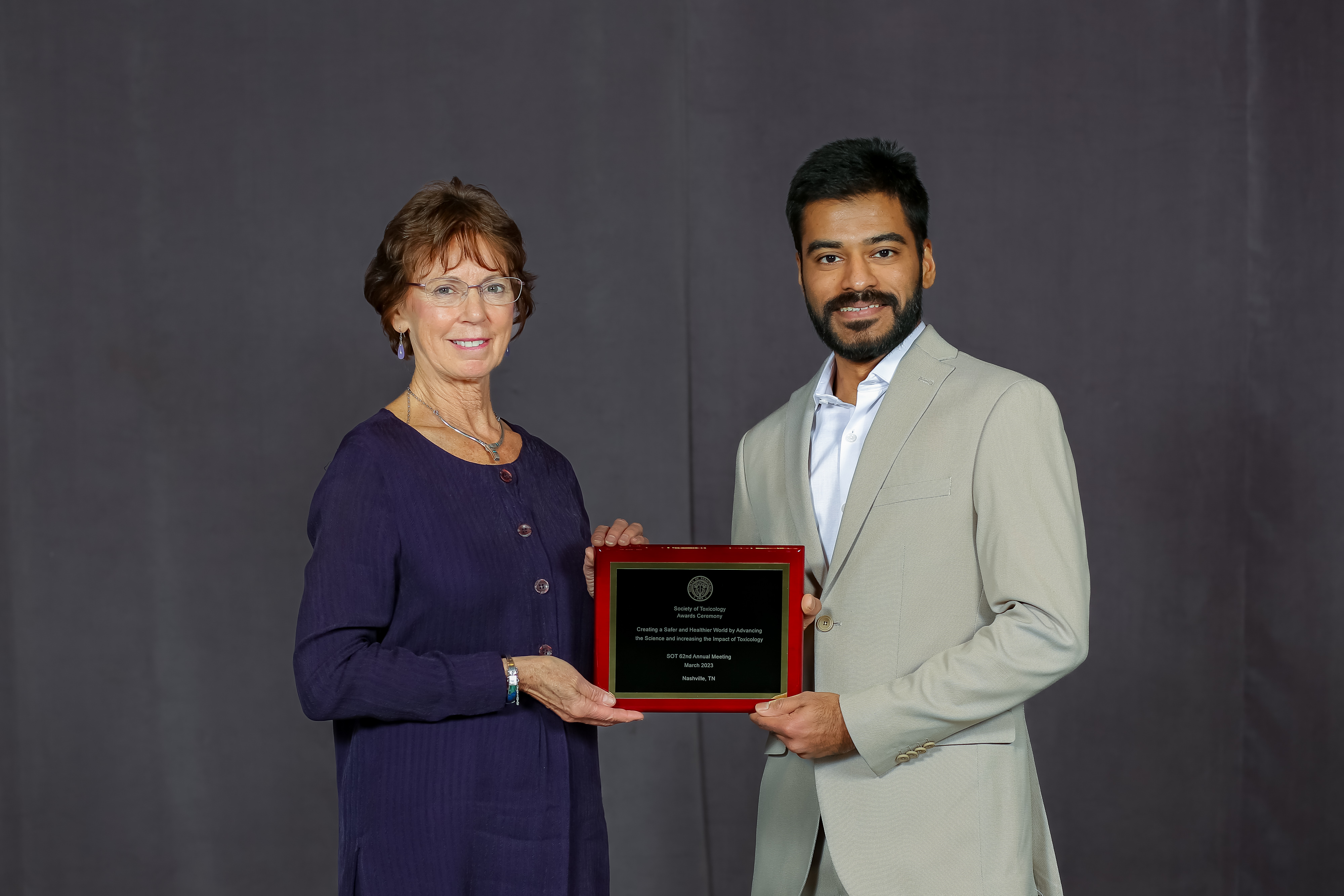 A woman is standing on the left and a man is standing on the right. Both are coming together to hold an award plaque while smiling.